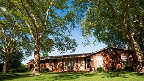 Une maison à deux étages, construite en briques, avec un porche à l’avant, entourée d’une pelouse bien entretenue et de grands arbres.