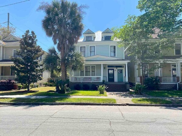 A two-story house with a front porch, a small front yard with a palm tree, and a clear blue sky. - Furnished Executive Rental in Victorian District! (Savannah)