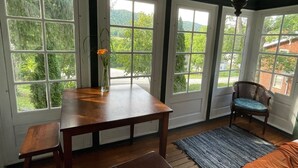 Une pièce avec une table en bois, deux chaises, un banc et un tapis. De grandes fenêtres offrent une vue sur la verdure et un bâtiment situé à l’extérieur.
