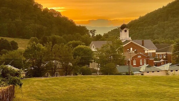 A rural landscape with a prominent building featuring a clock tower, surrounded by lush greenery and a sunset sky.