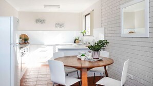 Una cocina moderna con armarios blancos, una mesa de comedor de madera y sillas blancas. Hay plantas en macetas sobre la encimera y una ventana sobre el fregadero.