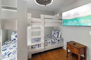 Una cama litera con ropa de cama temática náutica, una mesita de noche de madera y un televisor montado en la pared mostrando una escena de playa.
