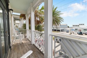 A covered porch with white railings, a wooden floor, and a view of the street with parked cars and buildings.