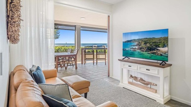 Un salon avec un canapé, un téléviseur posé sur un meuble et une vue sur la mer grâce à de grandes fenêtres.