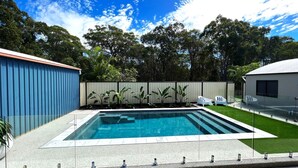 Una piscina rectangular con agua azul cristalina, rodeada por un patio de concreto. Hay sillas de descanso blancas y un pequeño césped con una cerca al fondo.