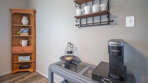 Uma cozinha compacta com cafeteira Keurig, um jarro de vidro e uma cafeteira sobre uma mesa. Há uma prateleira de madeira com xícaras e um vaso, e uma prateleira na parede com mais xícaras.