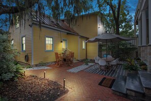 Una casa amarilla con patio, que incluye una mesa y sillas, rodeada de plantas y un sendero de ladrillos.