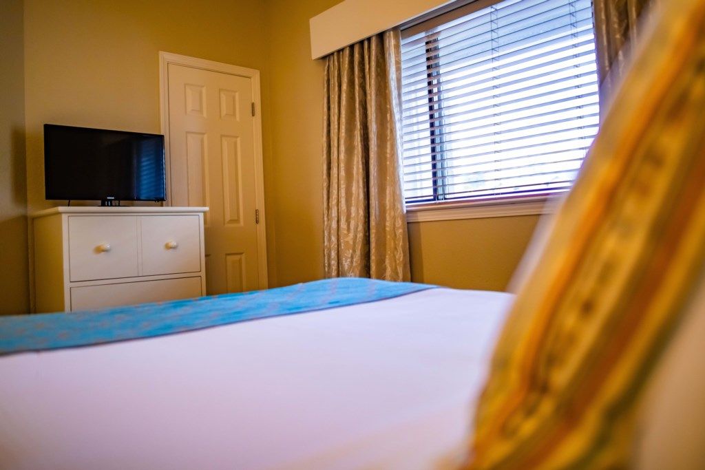 A bedroom with a bed, a TV on a dresser, a window with blinds, and a door.