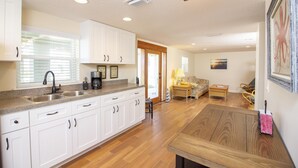 A modern kitchen with white cabinets, a double sink, and a wooden dining table. - Charming Cottage ~ Steps to the Beach ~ Pet Friendly (Saint Augustine)