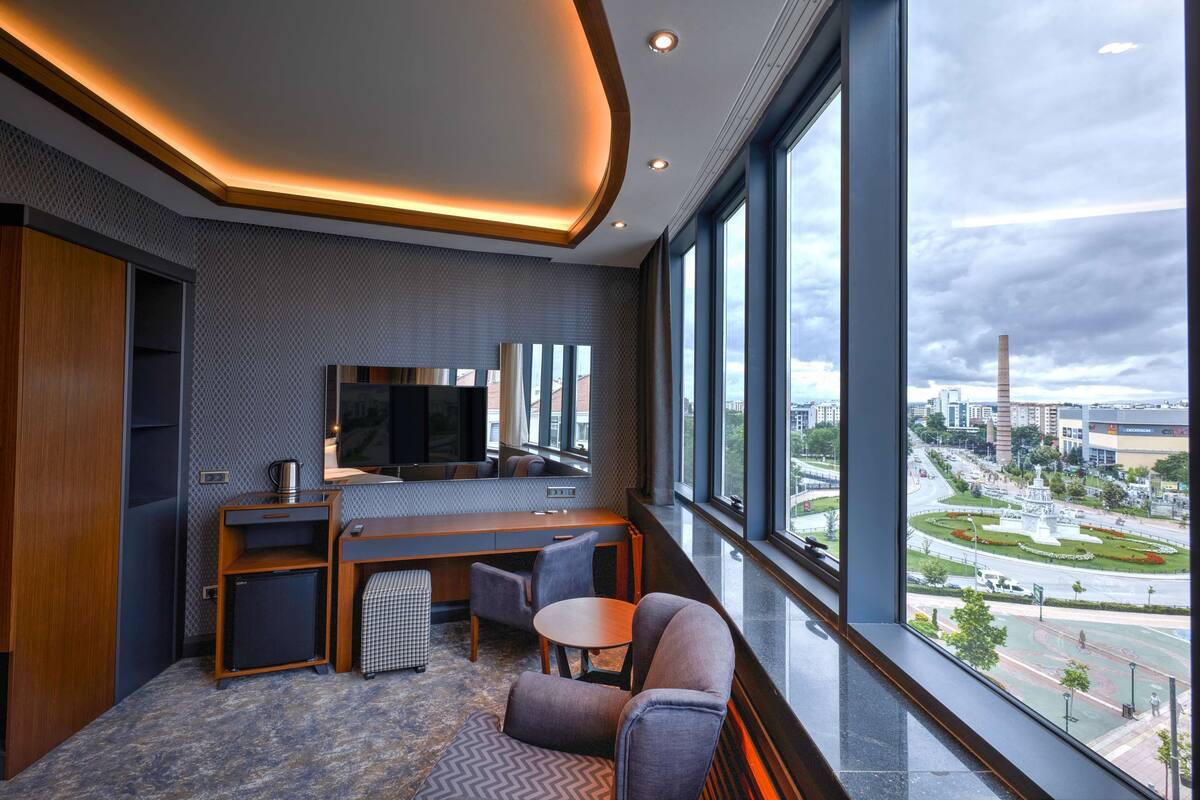 A modern hotel room with a large window, a flat-screen TV, a desk, and seating area.