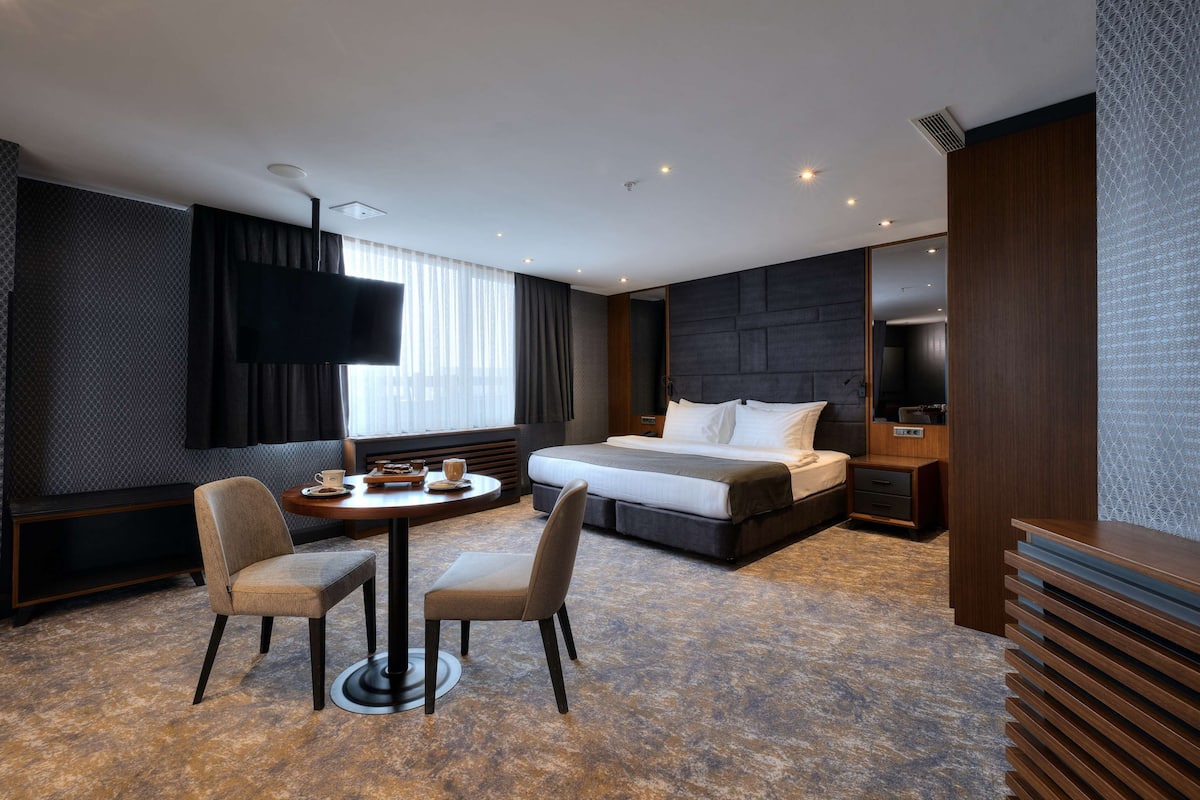 A modern hotel room with a large bed, a flat-screen TV, a desk with a chair, and a sofa.