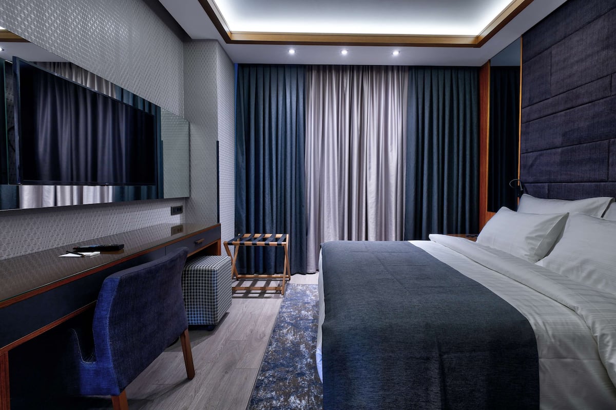 A modern hotel room with a large bed, a desk with a chair, a TV, and a large window with curtains.