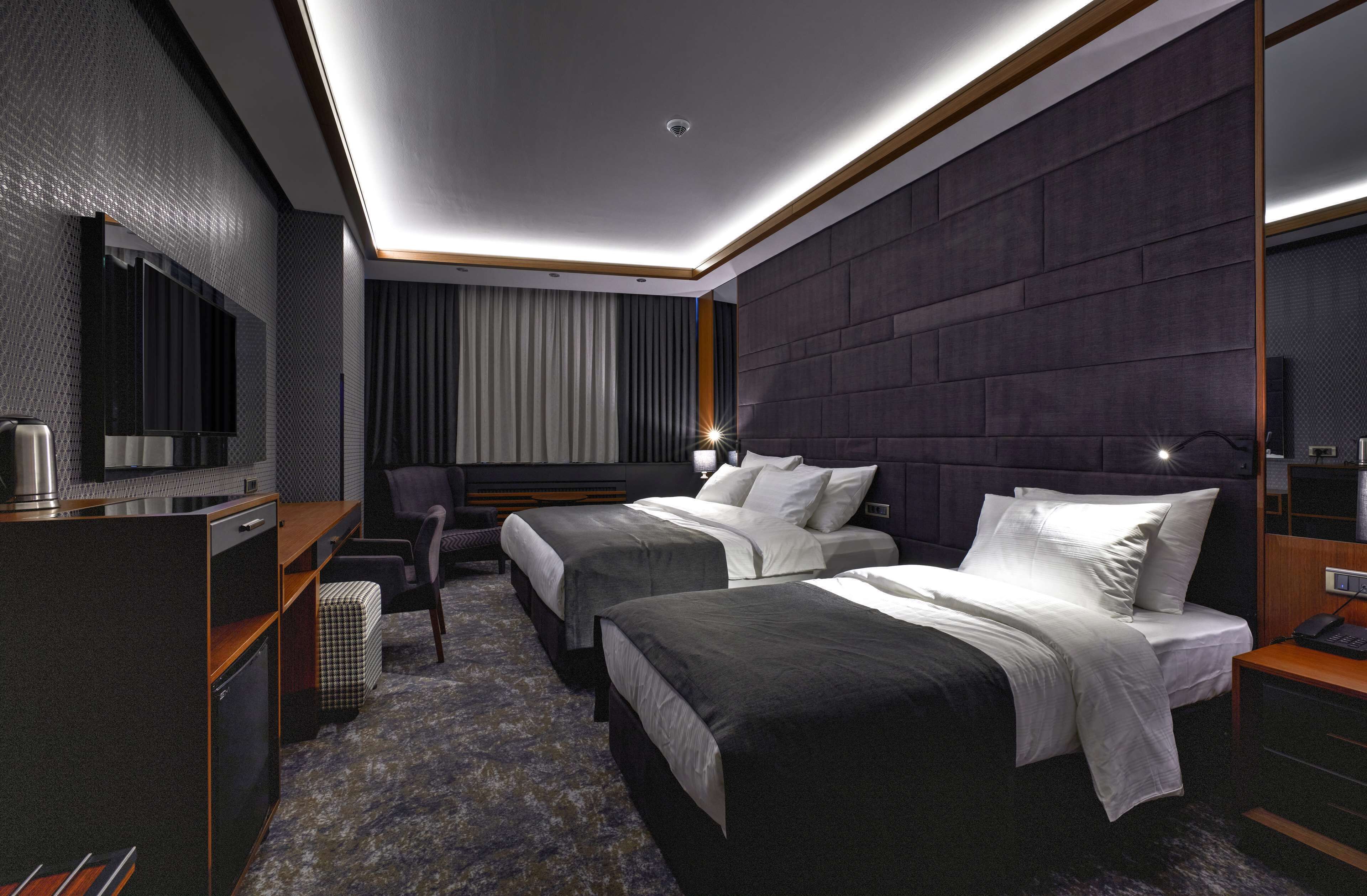 A modern hotel room with two beds, a desk with a chair, a television, and a large window with curtains.