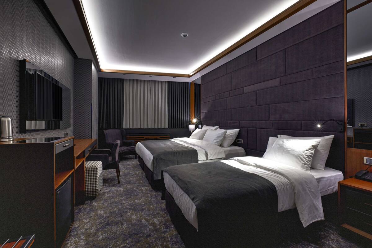 A modern hotel room with two beds, a desk with a chair, a television, and a large window with curtains.
