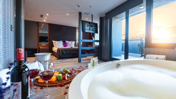 Une chambre d’hôtel moderne dotée d’une grande baignoire, d’un coin repas avec des fruits et du vin, et offrant une vue sur le coucher de soleil à travers de grandes fenêtres.