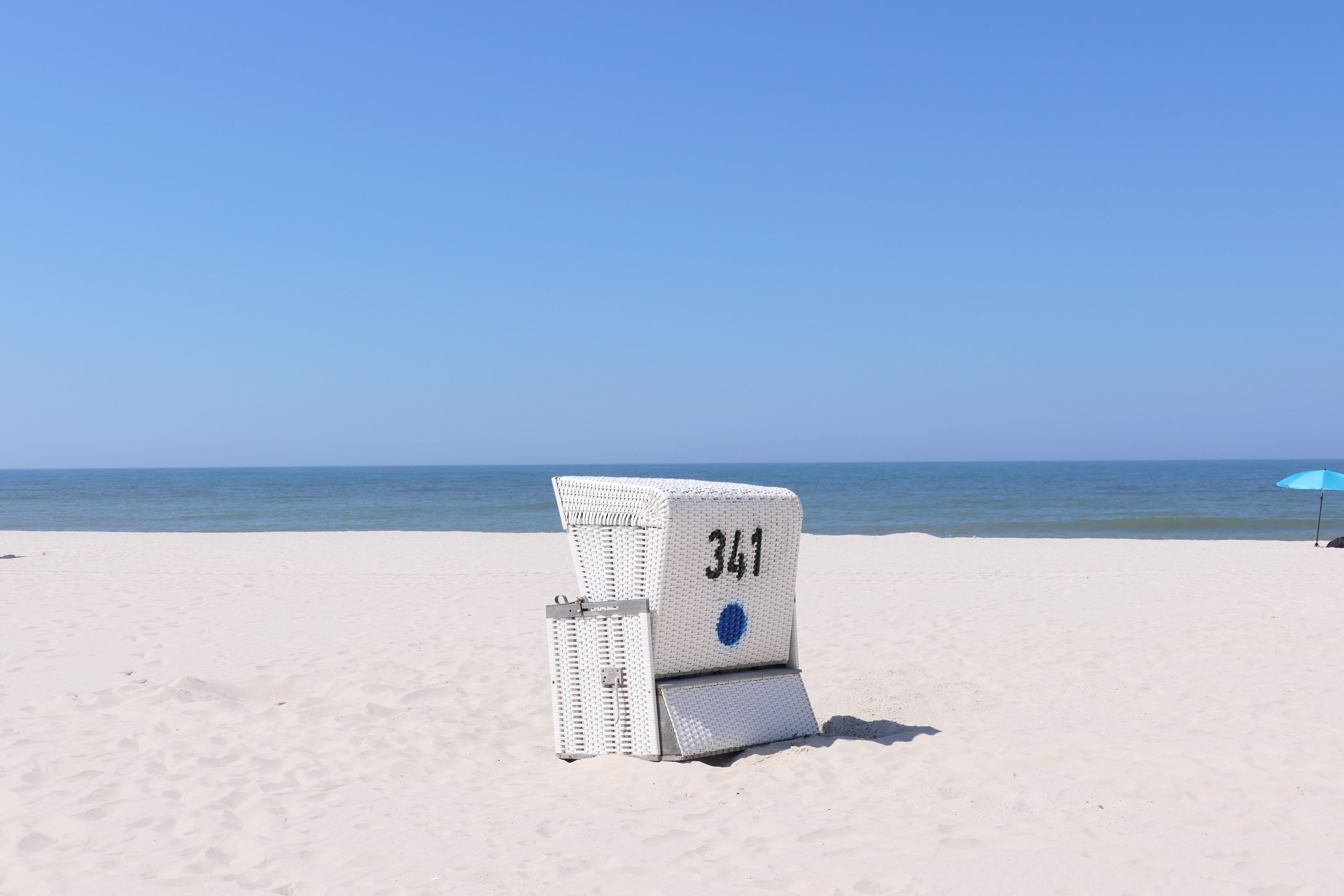 Una silla de playa con el número 341 sobre una playa arenosa, con un cielo azul despejado y el océano al fondo.