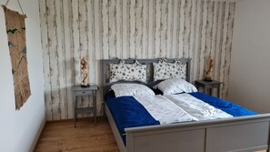 Un dormitorio con una cama, dos mesitas de noche, un macramé colgado en la pared y suelo de madera.