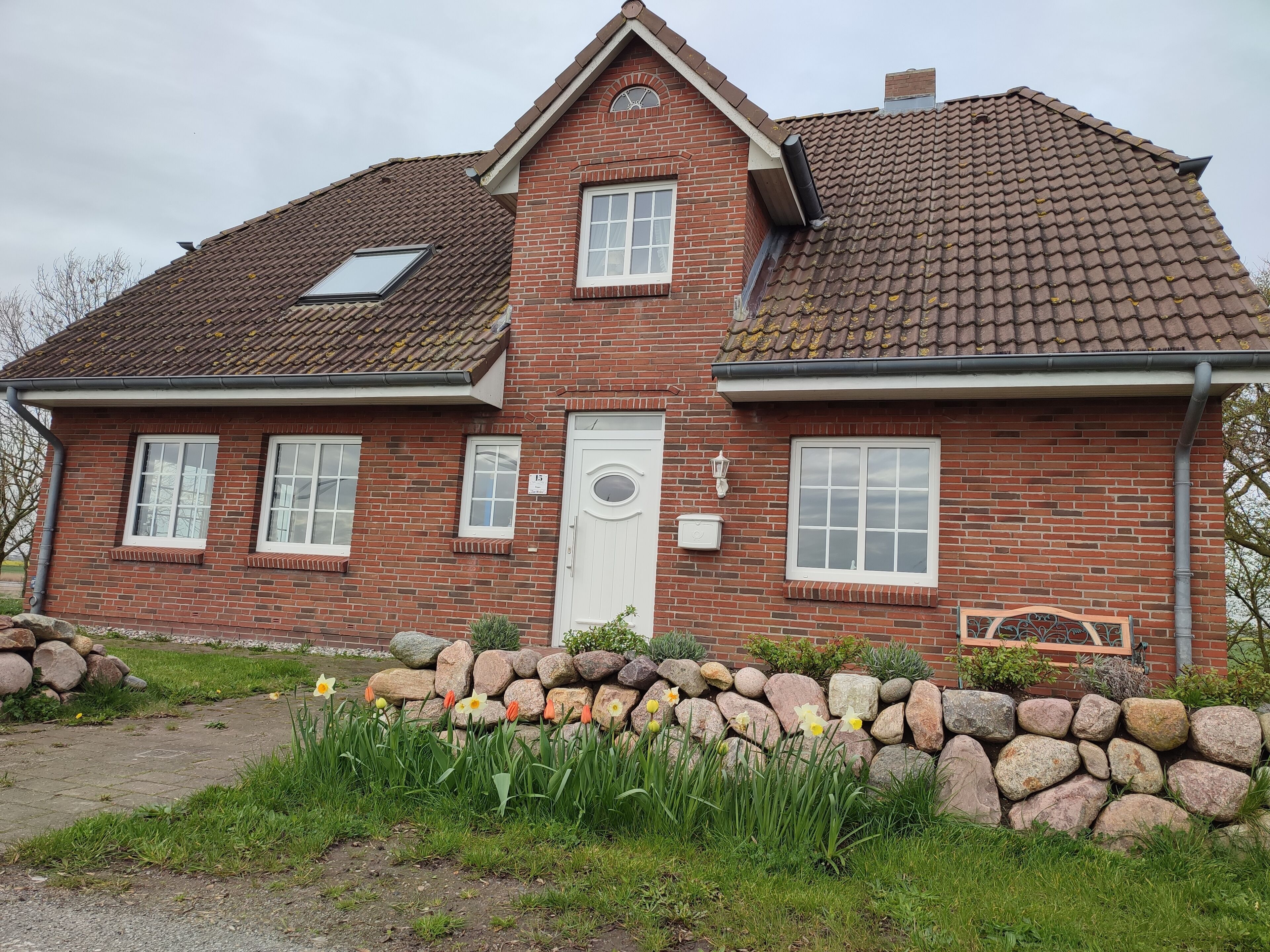 Une maison à deux étages, construite en briques, avec un toit en tuiles, une porte d’entrée et un petit jardin agrémenté d’un mur en pierre et de jonquilles.