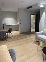Quarto de hotel moderno com piso de madeira, sofá cinza, mesa de centro redonda e uma mesinha de cabeceira. Há uma porta que dá para um banheiro com chuveiro e vaso sanitário.