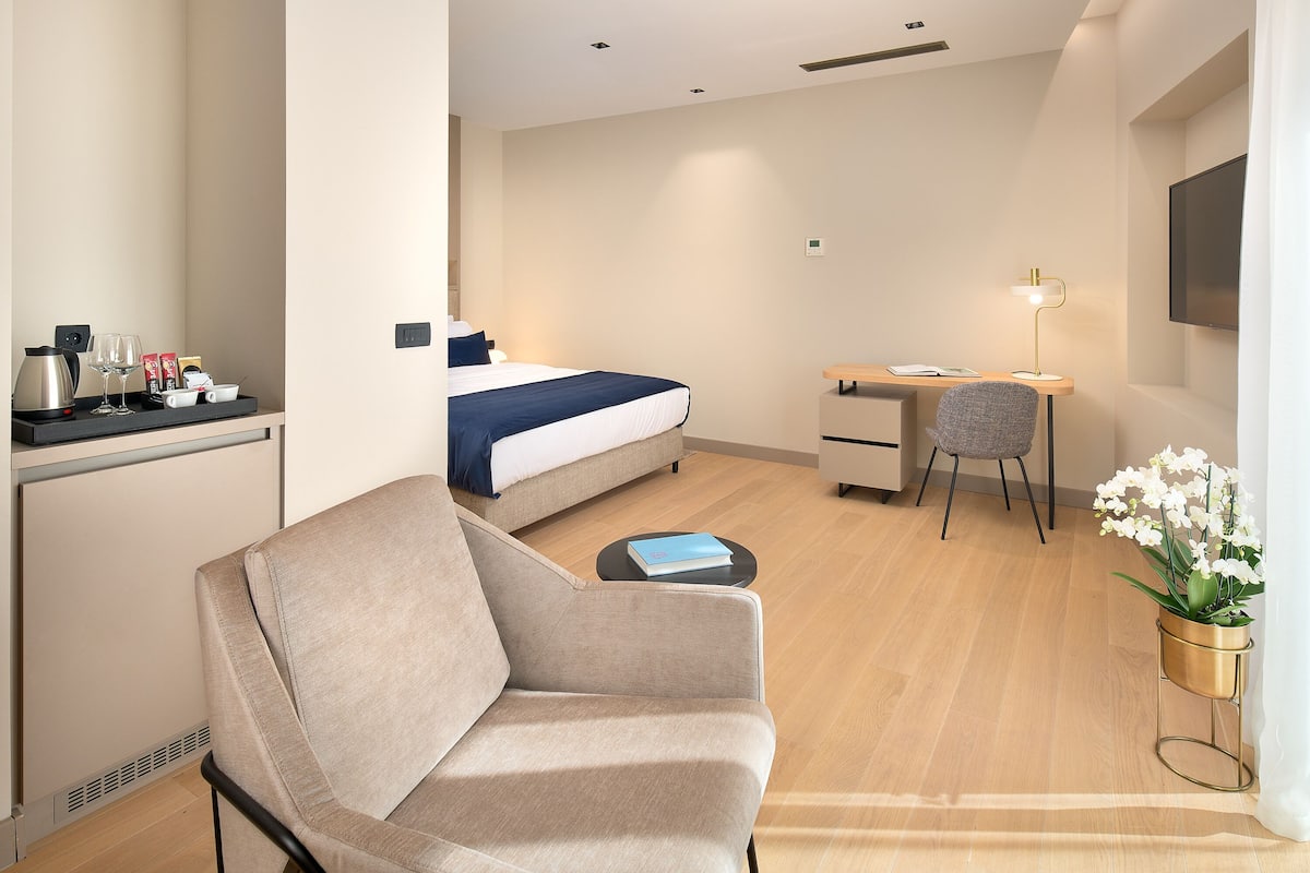 Ein modernes Hotelzimmer mit einem Bett, einem Schreibtisch, einem Stuhl, einer Couch, einem kleinen Tisch mit einem Buch, einer Kaffeemaschine und einer Topfpflanze.