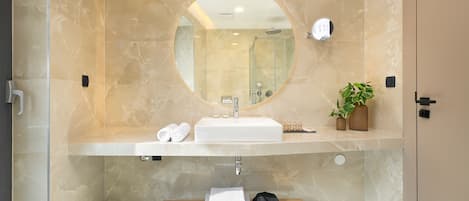 A modern bathroom with a marble-tiled wall, a round mirror, a sink, a towel rack, and a shelf with toiletries.