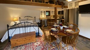 A bedroom with a bed, bedside table, and a nightstand. There is a wooden bench and a dining table with chairs. The room has a wooden ceiling with exposed beams and a chandelier.