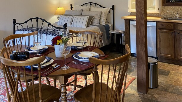 A dining area with a round table, chairs, and a chandelier. A bed with pillows and a nightstand with a lamp are visible in the background.