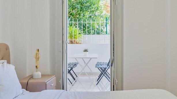 A bedroom with a bed, a bedside table with a lamp, and a view of an outdoor balcony with a table and chairs. - Garrett Terrace (Setúbal)