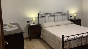 A bedroom with a bed, two bedside tables with lamps, a nightstand, a decorative glass dome on a table, and an air conditioning unit on the wall. - Villa Lupino (Orosei)