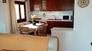 A cozy living room with a sofa and a dining area with a table and chairs. A kitchen with wooden cabinets and a microwave is visible in the background. - Villa Lupino (Orosei)