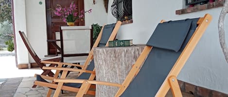 A covered outdoor seating area with wooden lounge chairs and a stone floor.