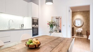 A modern kitchen with white cabinetry, a wooden dining table with a fruit bowl, and a view into a living area with a round mirror and a chair. - Regent Style Soho Loft - Best of Both Worlds (London)