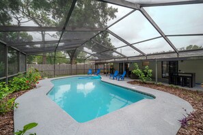 A covered swimming pool with blue chairs and a table, surrounded by plants and a wooden fence.