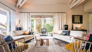 A cozy living room with a sofa, armchair, and coffee table, large windows with curtains, and a view of greenery outside.