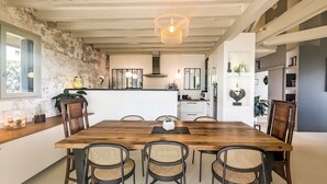 A dining area with a wooden table and rattan chairs, a chandelier, and a kitchen in the background.