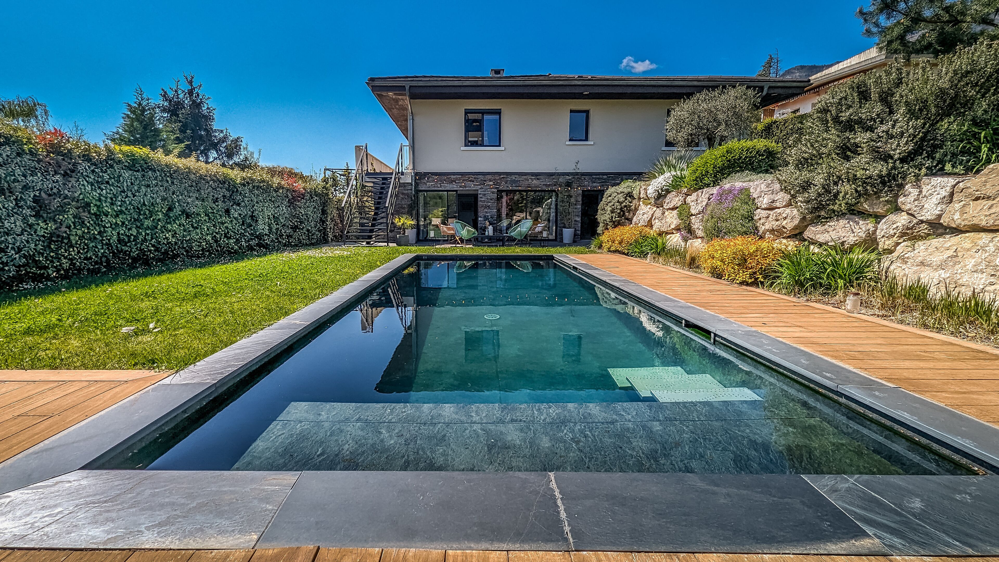 Une piscine rectangulaire dotée d’une terrasse en bois et de carreaux de pierre, entourée d’un jardin bien entretenu et d’une maison à la façade en pierre.