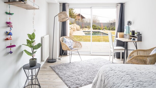 A modern bedroom with a large bed, a desk with a chair, a potted plant, and a view of a backyard with a pool.