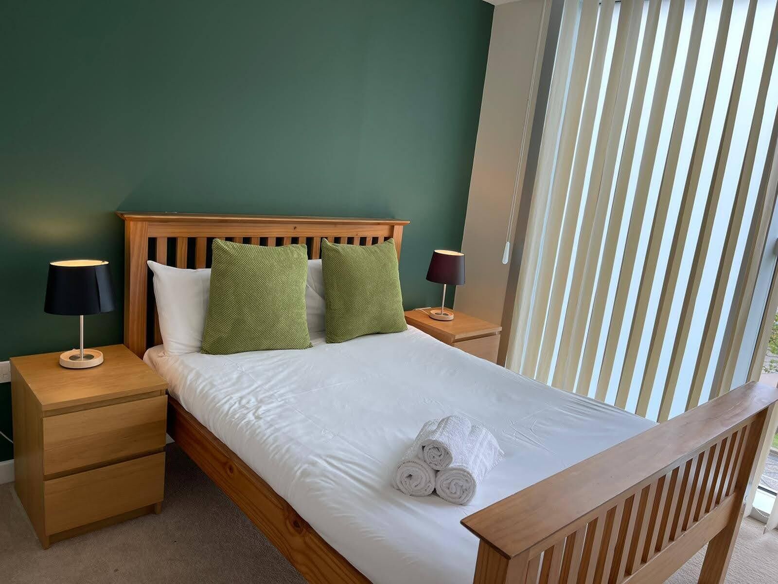 A neatly made bed with white linens and two green pillows, a wooden headboard, two bedside tables with lamps, and a window with vertical blinds.
