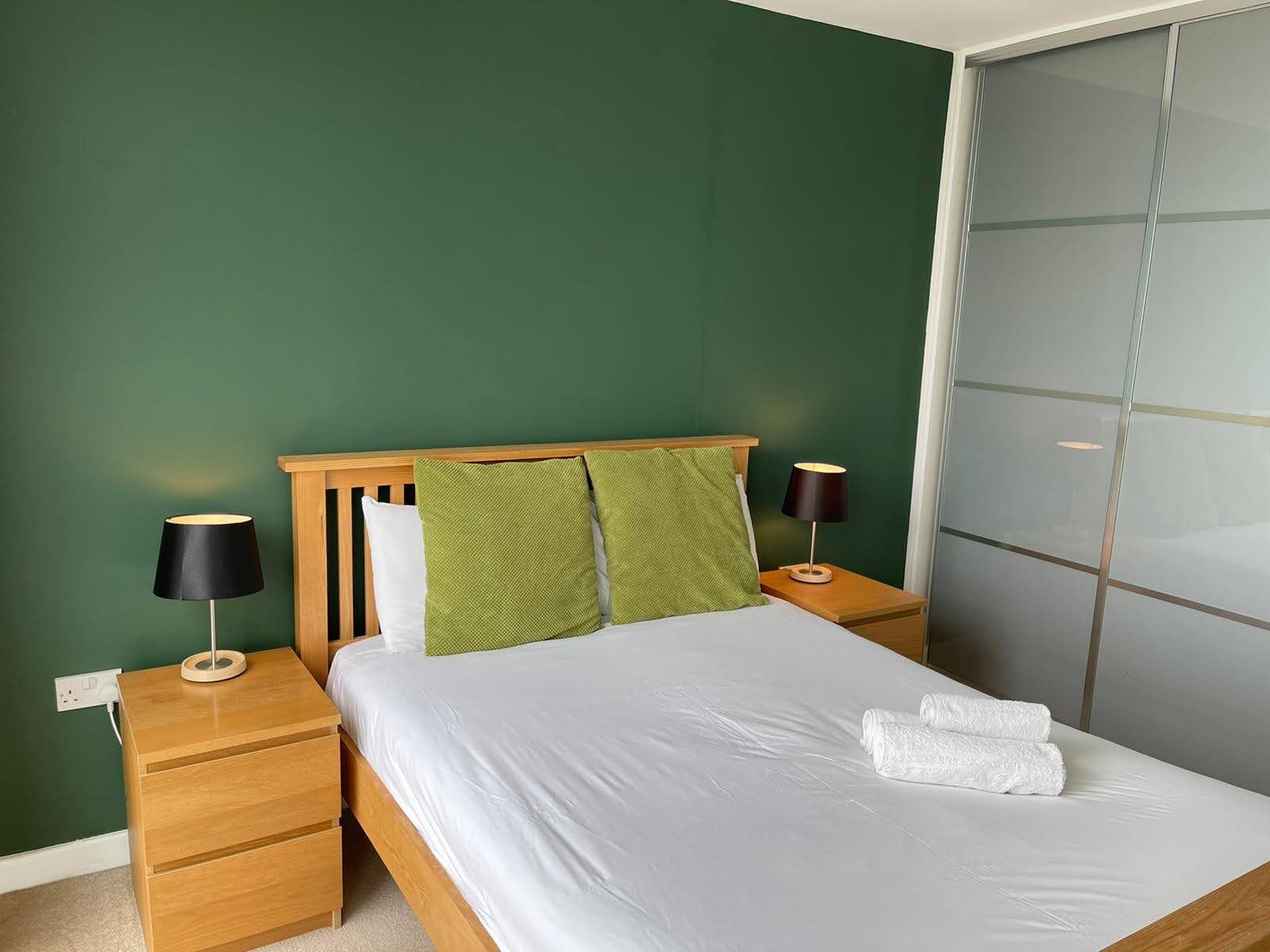 A neatly made bed with white linens, two bedside lamps, and a green pillow on a wooden headboard.