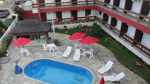A pool area with lounge chairs and umbrellas, surrounded by a brick patio and a building with balconies. - Pousada Lusitana (Ipojuca)