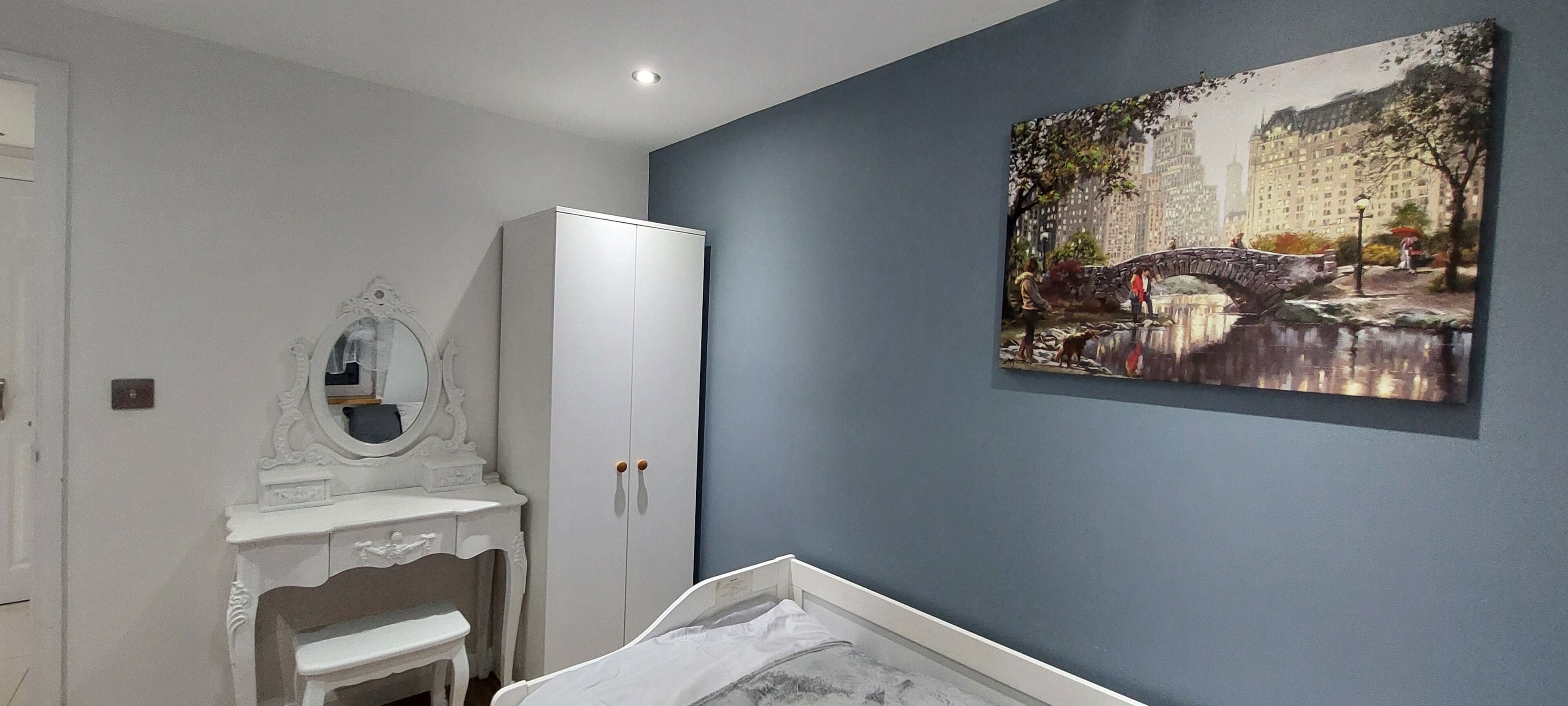 A bedroom with a bed, a white wardrobe, a vanity with a mirror, and a painting of a bridge.