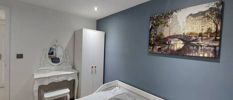 A bedroom with a bed, a white wardrobe, a vanity with a mirror, and a painting of a bridge.