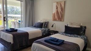 Une chambre d’hôtel avec deux lits, une grande fenêtre avec des rideaux, une petite table avec une lampe et un tableau au mur.