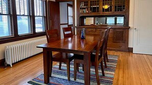 A dining room with a wooden table and chairs, a patterned rug, and a kitchen area with wooden cabinets and a window. - Classic Macalester Area St. Paul Duplex (Saint Paul)