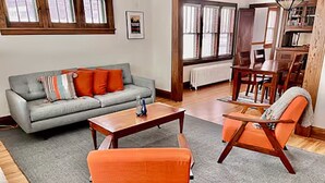A living room with a grey sofa, orange chairs, a wooden coffee table, and a dining area with a wooden table and chairs. - Classic Macalester Area St. Paul Duplex (Saint Paul)