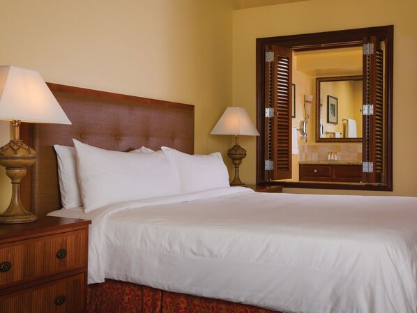 Une chambre d’hôtel avec un grand lit, deux tables de chevet avec des lampes, une tête de lit en bois, un grand miroir et une salle de bain visible à travers une porte ouverte.