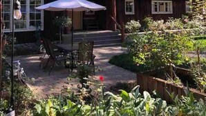 A wooden house with a patio area, surrounded by a garden and tall trees. - Cozy Historic Cabin Near Hiking (Wolf Creek)