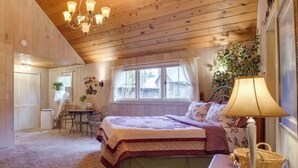 A cozy bedroom with a wooden ceiling, a bed with a floral bedspread, a desk with a chair, and a window with curtains. - Cozy Historic Cabin Near Hiking (Wolf Creek)