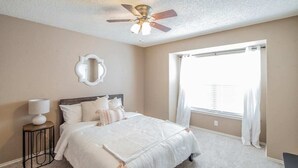 A bedroom with a bed, a ceiling fan, a window with curtains, and a wall-mounted mirror. - Fresh, Clean & Comfortable Place in Round Rock (Round Rock)