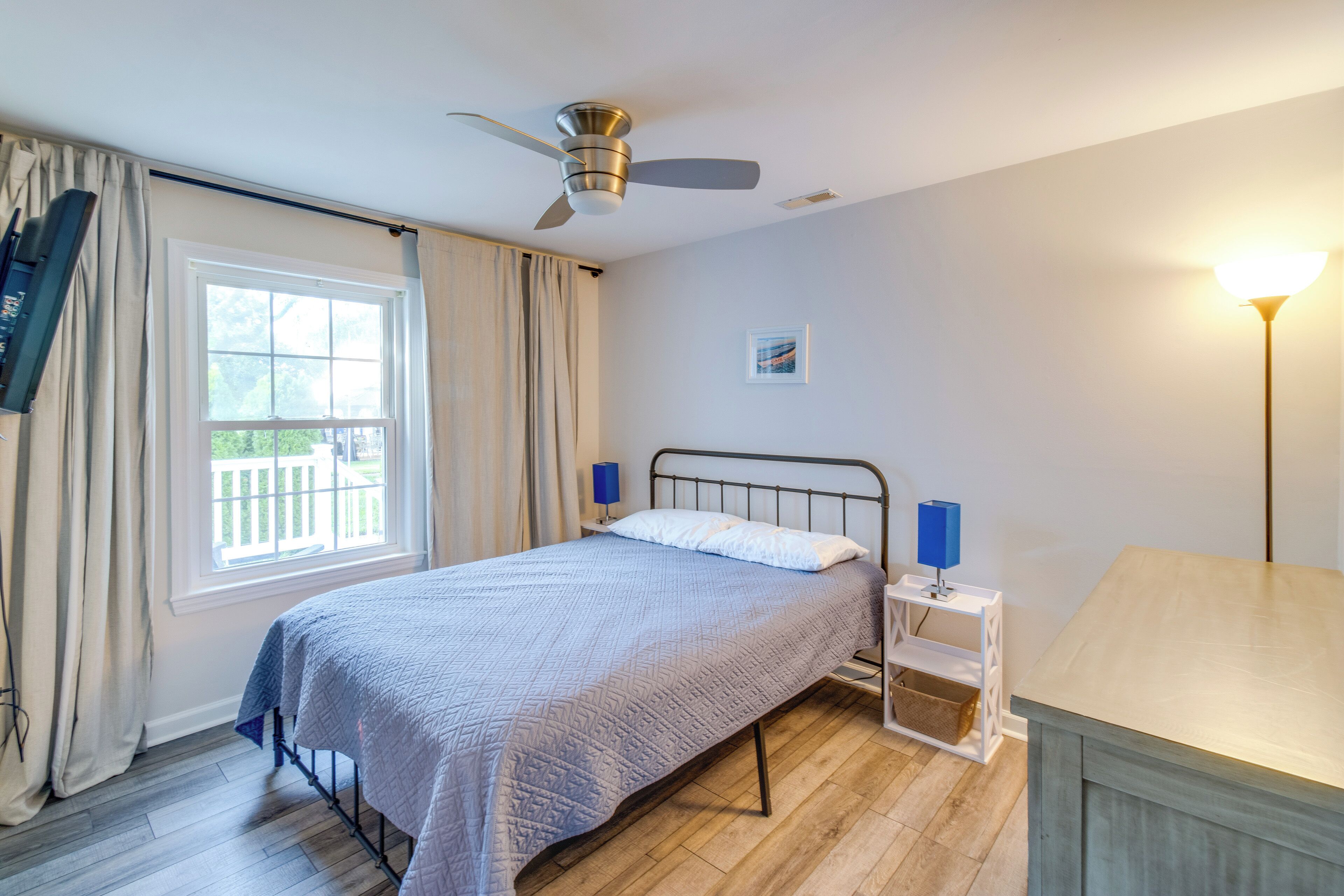 A bedroom with a bed, a ceiling fan, a bedside table with a lamp, a window with curtains, and a view of the outdoors.
